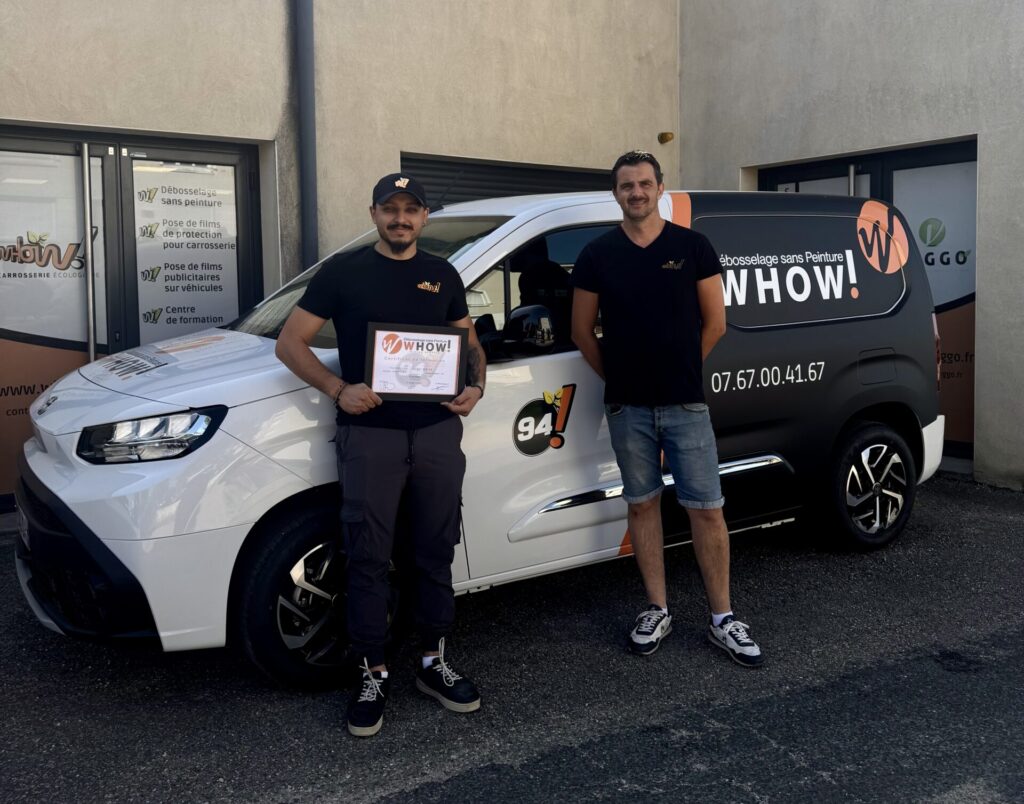 Deux hommes posent devant une voiture blanche avec un logo et un panneau de certificat.
