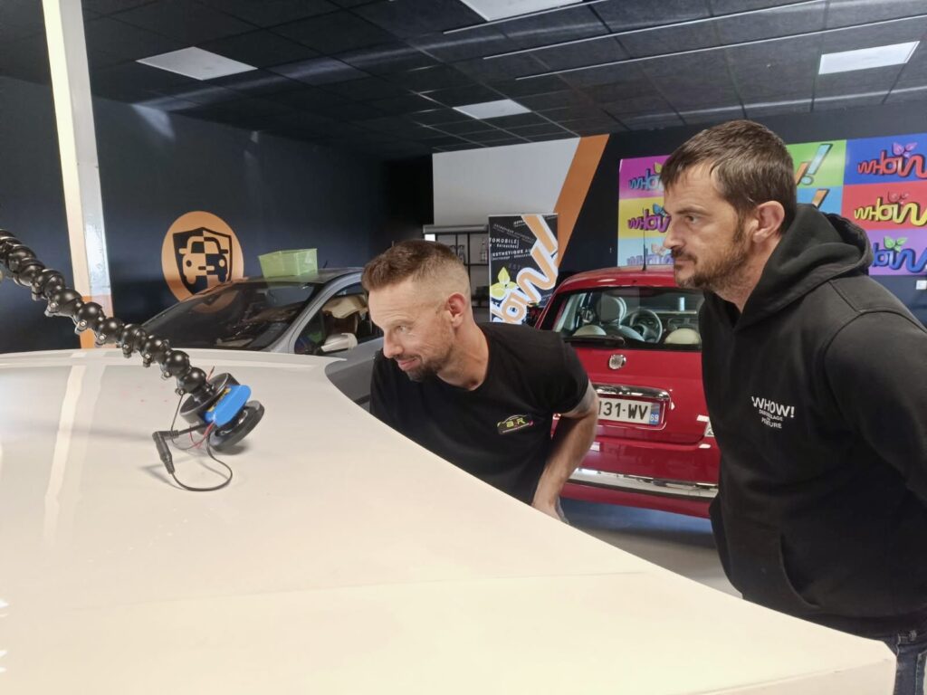 Deux hommes regardent une pièce de machine avec des voitures en arrière-plan